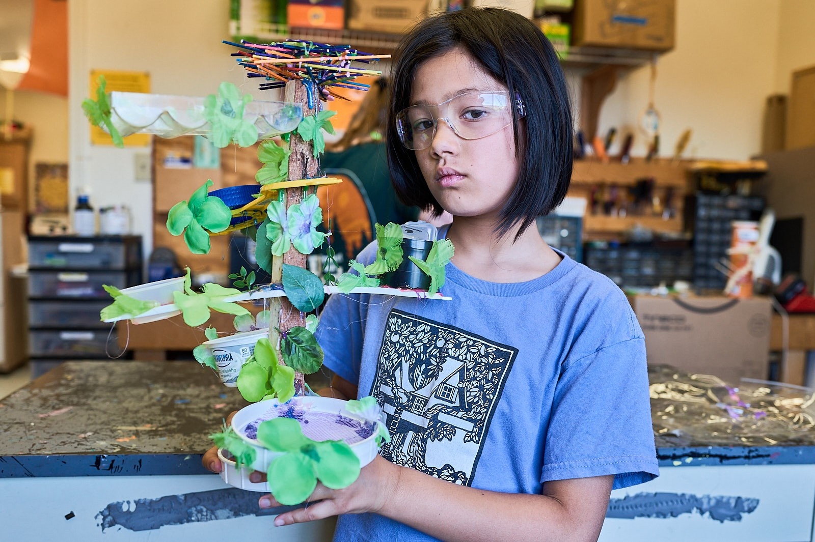 A young person with glasses and shoulder-length black hair stands in an art classroom, holding a creative sculptural project made with green materials, branches, and colorful elements.