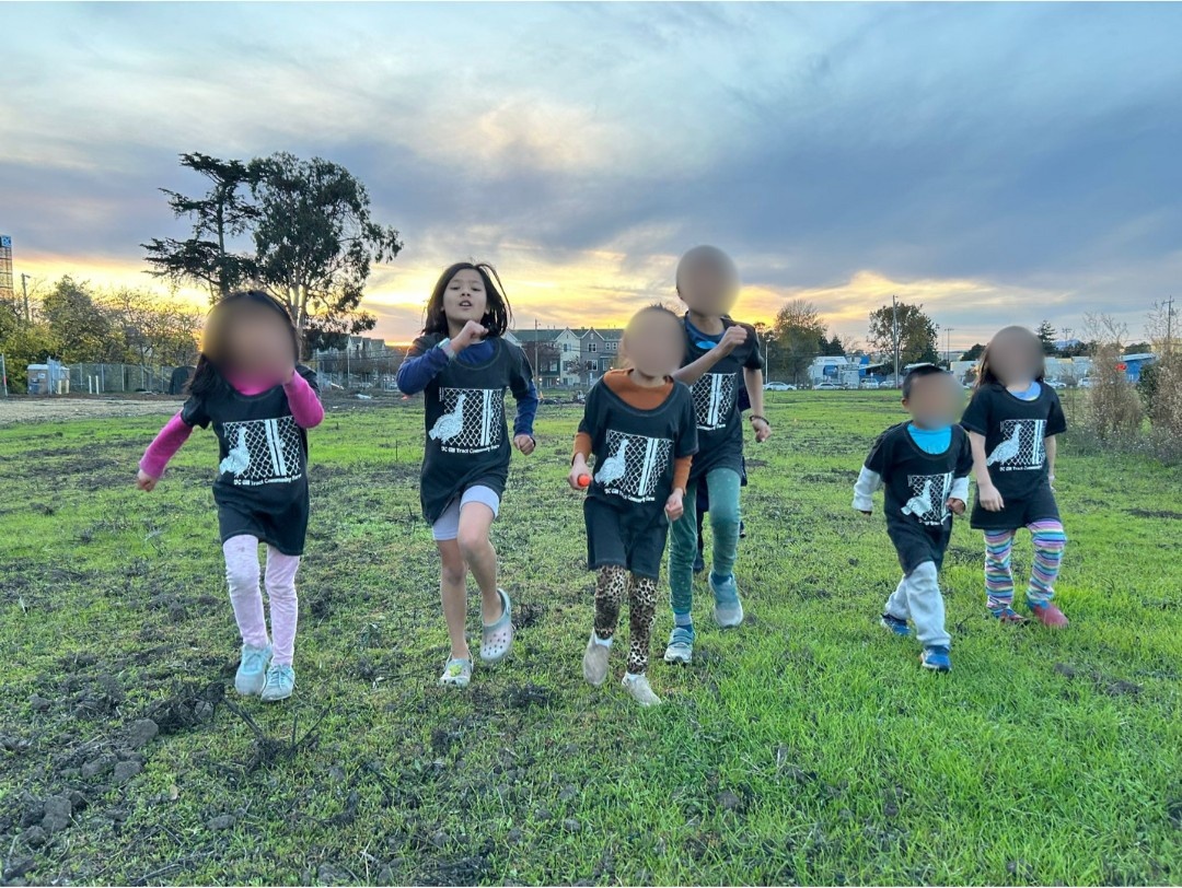 A group of children wearing matching dark shirts run together across a grassy field at sunset, with trees and buildings visible in the background