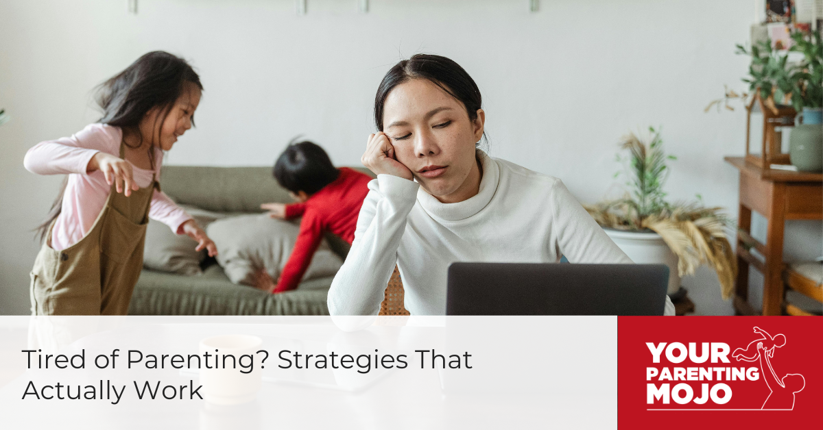 A tired parent with head in hand works on laptop while two children play energetically in the background on a couch